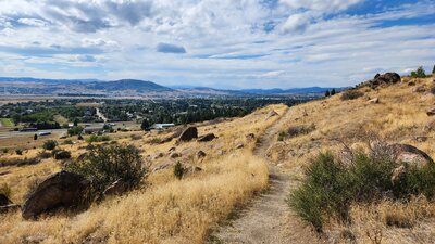 The mining heritage trail undulates across dry grassland with scattered rocks. The City of Butte is in the background.