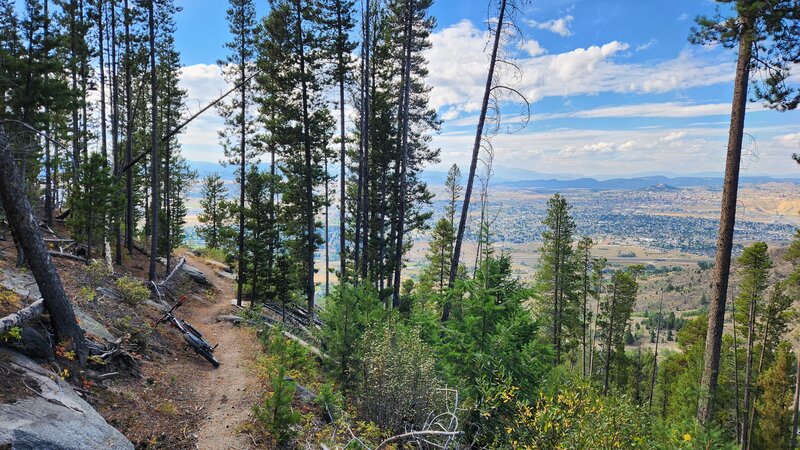 Narrow trail traverses a steep slope with views of the City of Butte in the background.