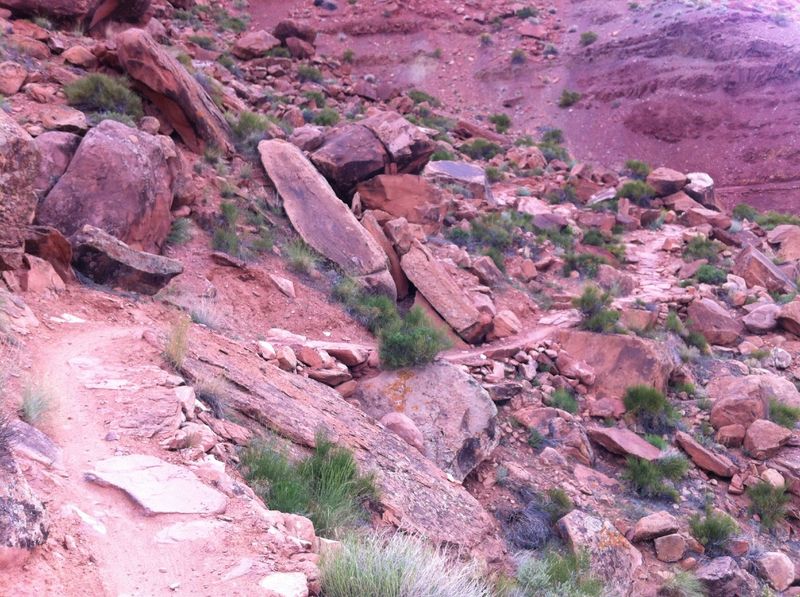 You can see one of the steep rock armored climbs. Photos can be deceiving. After another steep switchback the trail hooks around the rocky hill in the foreground and pops out on narrow bench cut sandstone dirt which can barely be seen in the background.